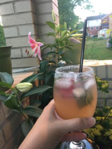 A hand holds a glass with a peach-coloured drink and ice cubes in it. The background is an image of a garden, the drink looks refreshing on a warm day.